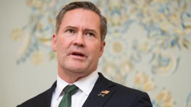 Mike Waltz stands in front of a wall that has a floral design. He is wearing a dark suit and a green tie.