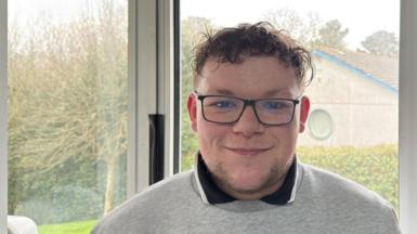 Dylan Caulkin, a young man with short curly hair in glasses, smiling wearing grey sweatshirt and collared T-shirt, standing in front of a window looking out on a hedge and tree