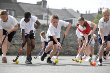 A row of pupils in white and black sports PE kits carefully guiding small balls with hockey sticks with a grey stone wall in the background