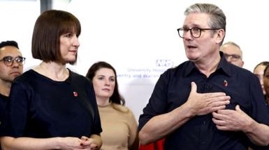 Chancellor of the Exchequer Rachel Reeves on a hospital visit with Prime Minister Sir Keir Starmer. Both are wearing navy clothes and small poppy badges, and smiling, and there is a team of medical staff behind them