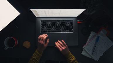 Aerial view of hands typing on a laptop, with a cup of tea, a biscuit, and notepads filled with notes on a dark desk.