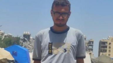 Imad Qudaih stands in front of destroyed buildings and a bright blue tent, wearing a light blue t-shirt and black glasses.