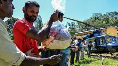 A man carrying a plastic bag full of food, handing it to the man beside him. They're part of a line helping to carry food out of a helicopter.