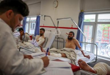 A doctor takes notes at a hospital in Kabul, where three men sit in hospital beds. Two of them have bandages - one to a leg, and another to his arm. The bandaged leg of a fourth men can be seen in the side of the shot.
