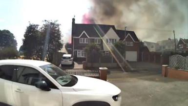 Smoke and flames are rising above a house with a white car parked on the drive.