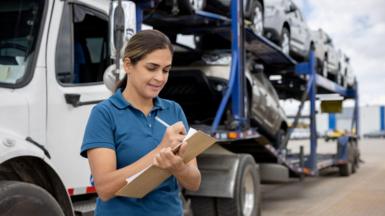 A woman with long straight brown hair in a ponytail wearing a blue uniform style polo shirt writing on a clipboard in front of a lorry full of cars