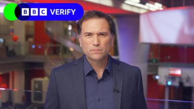 Ros Atkins in a dark jacket and dark shirt looks directly at the camera, with a number of screens and the red walls of the BBC newsroom visible behind him.