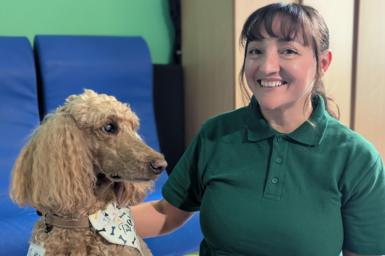 Katia Stanford, a woman with dark hair tied back and a fringe, wearing a dark green polo shirt, smiles at the camera with her hand on a biscuit-coloured poodle.