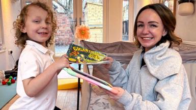 Romi, who has blond curly hair and is wearning a white t-shirt, is passing a book to his mum, Alexis who has short brown hair and is wearning a grey hoodie. She is passing a digital device with a colourful cover back to him and both are smiling. 