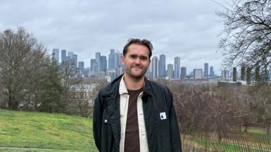 A young white man with brown hair, dressed in brown tshirt, white shirt and black jacket smiles at the camera with a backdrop of trees and skyscrapers