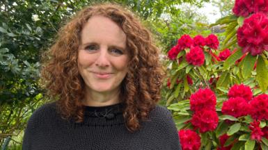 Maggie O'Farrell with curly auburn hair in black jumper. Background shows leaves on left and red flowers on the right