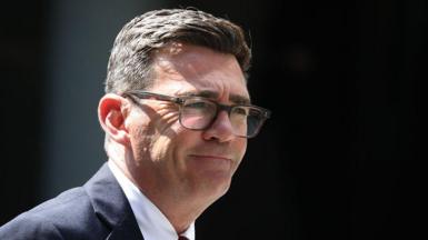 Andy Burnham is wearing glasses, a dark-coloured suit jacket, white shirt and a tie. He has a short, dark hair.