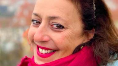 A brunette woman with brown hair, a pink scarf and pink lipstick. She is smiling at the camera