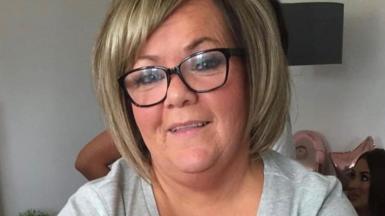 Susan McGowan looks into the camera smiling - she has blonde hair in a short bob, black-rimmed glasses and a light grey t-shirt