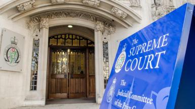 Entrance to the Supreme Court in London, a white brick building covered in ornate carvings, including a big blue sign reading "the Supreme Court"