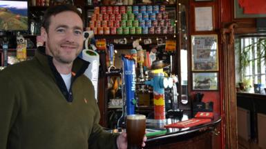 A man in a khaki fleece is holding a pint of beer and smiling at the camera. He is stood in front of a bar that has bar taps, tubs of Pringles and posters on the wall