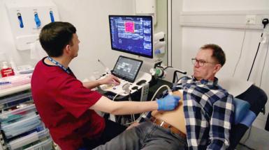 Phil Brown, who has brown hair and glasses, lying down wearing grey jeans and a blue checked shirt that is open at the waist. A technician wearing red scrubs and blue gloves holds the ultrasound device to Phil's stomach while he examines the results on screen