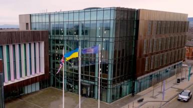 Redcar and Cleveland Council's headquarters photographed from high up. There are three, interlinked square brown buildings. They are clad with the two on either side containing large glass frontages. The middle building, which sits back slightly from the other two, has a large area of wood-effect cladding with windows in the front separated by long, thin white pillars. In front stand three large flagpoles flying the flags of, left to right, the United Kingdom, Ukraine and the council's crest in gold on a blue background.