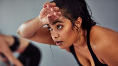 A close up shot of a woman wiping sweat from her brow as she works out, another out-of-focus person can be seen in the background.