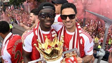 Bukayo Saka and Mikel Arteta, both in sunglasses and red and white team kit, with the Premier League trophy on Arsenal's bus parade