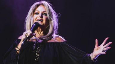 A glamorous woman with blonde hair, wearing all black and holding a microphone, with one hand raised up in the air.