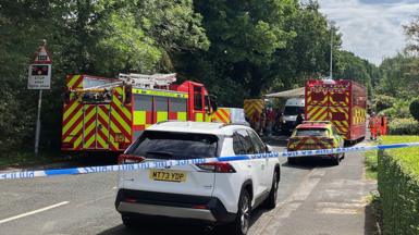 A fire engine is parked on the left side of a road with a fire and rescue car on the other side. There is a truck with Command Unit written on the back in the background and police tape is cordoning off the scene.