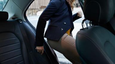 A woman stepping in to the back seat of a car from a city street, wearing a navy blazer and beige trousers