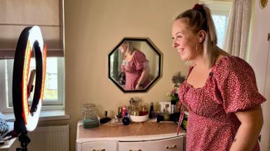 Roxanne stands in front of her light talking into her camera on her phone wearing a dress