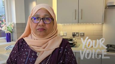 A woman in a peach hijab and glasses stands in a modern grey-hued kitchen. 