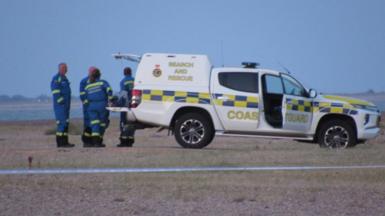 A white Coastguard vehicle with the words "Search and Rescue" and "Coastguard" printed on the side. It is on a beach and beyond it is sea. There are four people in blue boiler suits at its rear door, which is open.
