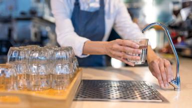 Stock photo shows a person filling a glass of water from a filter tap in a hospitality setting - they are positioned behind a bar with a stack of glasses in frame and wear a blue apron.