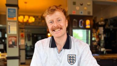 Sam O'Brien West, wearing an England football T-shirt, smiles into the camera while sitting at a table inside a pub