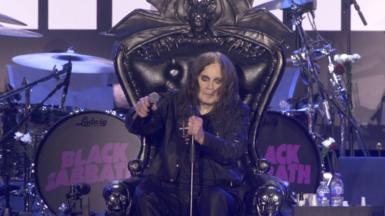 Ozzy Osbourne, who wears all black and has long brown hair and heavy eyeliner, sits on a throne onstage