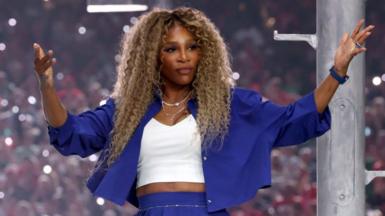 Serena Williams wearing a blue jacket and white t-shirt is holding up her arms