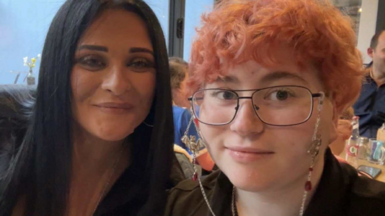 Two women side by side, looking at the camera and smiling. The woman on the right has short dyed-pink hair, glasses and is wearing a black top. The woman on the left has long dark brown hair and is wearing a black top. Behind them are tables with other people sat at them.