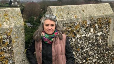 Monique Bonney in front of a stone wall. Behind her is a view of north Kent, including fields, a house and in the distance a town.