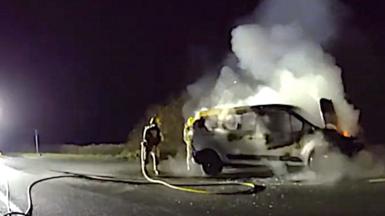 A burning van with a firefighter standing next to it with a hose.