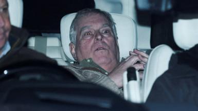 A horizontal grey-haired man looking alarmed in the back seat of a car
