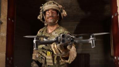 A military drone operator in camouflaged combat fatigues and helmet holds a drone with four propeller arms in his upturned, outstretched palm. He's standing inside the doorway of bare, concrete shelter.