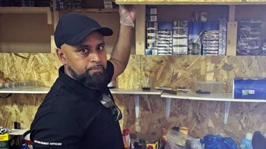 A person wearing a black shirt with 'ENFORCEMENT OFFICER' written on the sleeve is standing in front of wooden shelves. The person is lifting a wooden panel to reveal several stacks of boxes inside the shelf. Various items, including plastic containers, cleaning supplies, and other miscellaneous objects, are placed on the shelves below. A dog with black and white fur is visible at the bottom right corner of the image