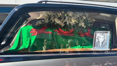 Close up of Bonnie Tylers coffin, which is covered in a Welsh flag. There is a black and white photo of Bonnie Tyler next to the coffin in a silver frame.