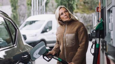 A stock photo of lady in a brown jacket filling up her car with fuel at a petrol station.