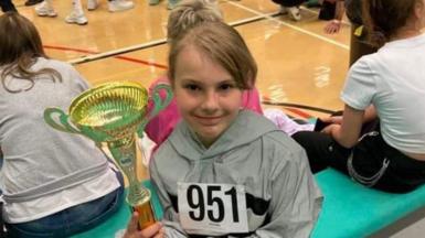 A young girl, her hair tied back, sits on a bench with other girls similar in age to herself. She is holding a trophy.