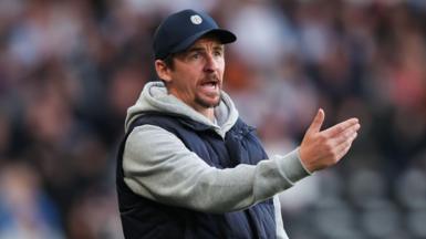Joey Barton, who is wearing a grey hoodie and gillet, gesticulates from the touchline at a football match. 