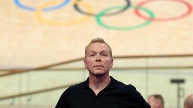 Sir Chris Hoyle, looking calm in a black top, stands below the Olympic rings