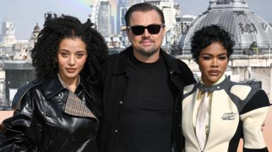 Chase Infiniti, Leonardo DiCaprio and Teyana Taylor at a promo event in London last year. Infiniti is wearing a black PVC jacket, DiCaprio is wearing a black T-shirt and jacket with sunglasses, while Taylor has on a racing-style cream and black zipped jacket.