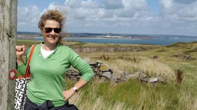 Clare Lane smiling while standing on a coastal path on a sunny day.  Clare is wearing a green sweater, white t-shirt and sunglasses. She has light brown curly hair that is blowing in the breeze.  