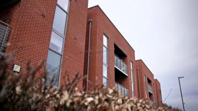 FA three storey, red brick block of flats with balconies enclosed by glass and steel barriers. A slate grey sky. 