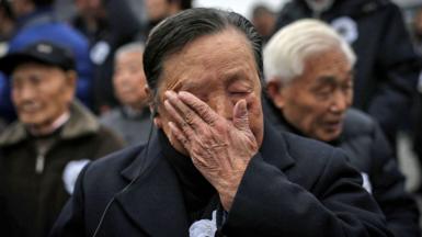 An elderly man wearing a suit with an ear piece in his ear wipes tears off his face. Behind him more elderly men can be seen, slightly blurred