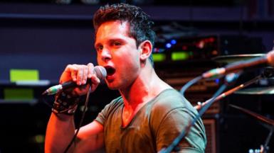 Man with brown hair sings into microphone on stage with red lighting shining down on him. He wears a tan t-shirt and black punk wristband.